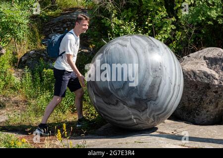 Mann posiert mit Steinglobus, Teil der Pars Pro Toto Skulptur von Alicja Kwade, auf der Helsinki Biennale 2021 auf der Insel Vallisaari in Helsinki, Finnland Stockfoto