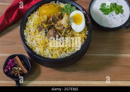 Köstliche Hammelfleisch Biryani serviert in schwarzem Topf mit Raita. kalkutta Stil Dum Biryani mit Hammelfleisch, gekochtem Ei und Kartoffeln. Holzhintergrund. Traditionelle m Stockfoto