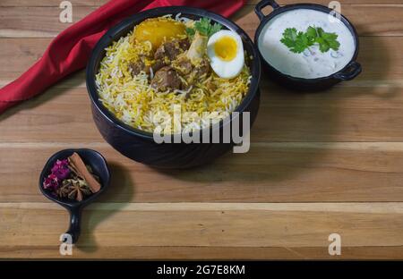 Köstliche Hammelfleisch Biryani serviert in schwarzem Topf mit Raita. kalkutta Stil Dum Biryani mit Hammelfleisch, gekochtem Ei und Kartoffeln. Holzhintergrund. Traditionelle m Stockfoto