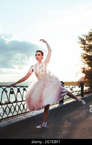 Eine dünne Ballerina in einem pinken Seiden-Korsett-Kleid hebt ihr bein mit spitzenzägen anmutig an den Flussufer in einem gusseisernen Zaun in der Stadt zurück Stockfoto