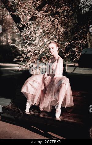 Im Sommer sitzt eine Ballerina in einem langen rosa Kleid auf der Rückseite einer Bank mit weit auseinander gespreizten Beinen und ihren Füßen in spitzenschuhen auf Zehenspitzen Stockfoto