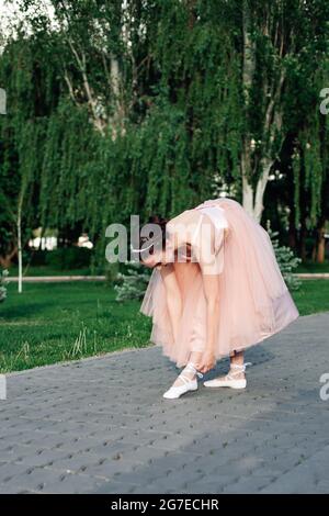 Lifestyle-Porträt einer Ballerina in einem rosa Kleid mit vollem Organza-Rock, das sich bis zum Bein beugt, um Bänder auf spitzenschuhen oder Ballett zu binden Stockfoto