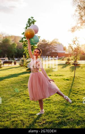 Porträt einer schlanken Ballerina in einem rosa Seidenkleid und weißen spitzenschuhen, stehend und balancierend auf einer Hand mit Luftballons in ihren Händen über ihr Stockfoto