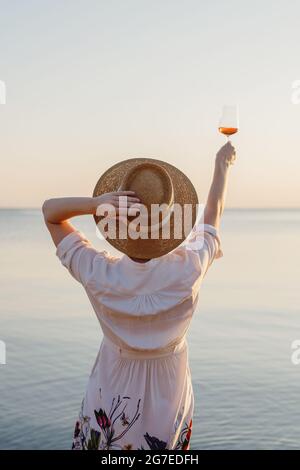 Rückansicht einer schönen Frau im Strohhut, die ein Glas Wein mit Meer im Hintergrund genießt. Stockfoto