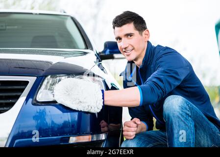 Lächelnder Mann, der die Stirnlampe seines Autos putzt und sie mit einem Handschuh wischt, während er sich neben dem Fahrzeug hockt Stockfoto