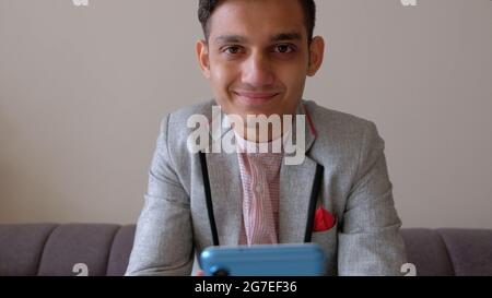 Porträt eines jungen lächelnden indischen Mannes in einer eleganten Jacke, der auf der Couch sitzt Stockfoto