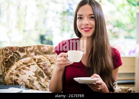 Hübsche Frau mit einem schönen Lächeln, die sich zu Hause auf einem Sessel entspannt und Kaffee oder Tee aus einer handgeführten Tasse und Untertasse trinkt Stockfoto