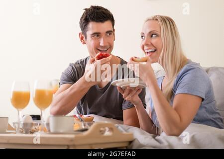 Ein fröhliches, verspieltes Paar genießt das Frühstück im Bett und grinst sich gegenseitig an, während sie sich darauf vorbereiten, Toast und Marmelade mit einem Aufstrich Orangensaft und Kaffee zu beißen Stockfoto