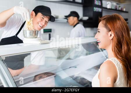 Lachender junger asiatischer Mann, der mit einem freundlichen Lächeln eine hübsche Frau mit Käsewägen an einer Feinkosttheke servierte Stockfoto