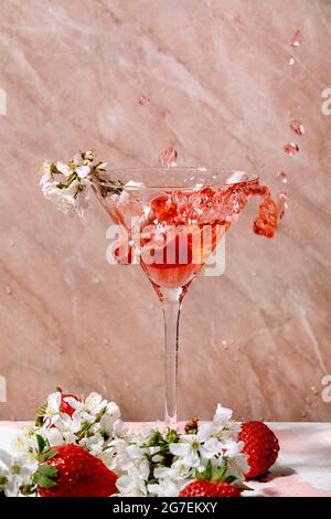 Spritzer alkoholischer oder alkoholfreier Erdbeer-Cocktail aus martiniglas, verziert mit blühenden Kirschbaumbästen über weißer und rosa Textur Stockfoto