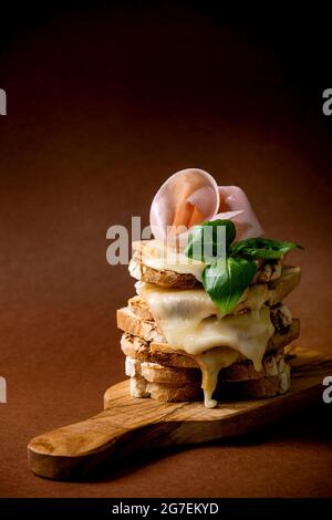 Vorrat aus gerösteten, geschmolzenen, Käse gepressten Sandwiches mit Schinkenfleisch, Basilikumblättern auf Holzschneidebrett auf dunkelbraunem Hintergrund Stockfoto