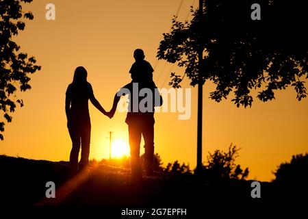 Familie bei einem Spaziergang bei Sonnenuntergang, das Kind sitzt auf den Schultern seines Vaters; die ganze Szene ist beleuchtet, sehr ruhig und friedlich Stockfoto