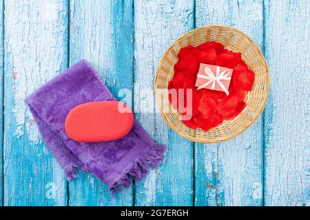 Auf einigen alten Holzbrettern in weißen und bläulichen Farben befindet sich ein fliederfarbenes Handtuch, auf dem sich ein rosa Schwamm befindet. Daneben ist ein Korb mit roten Blütenblättern und Stockfoto