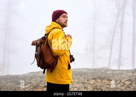 Junger männlicher Reisender in gelbem Mantel und Hut mit Kamera zum Entspannen im Freien bei nebligen Wetter. Urlaub und Lifestyle Wanderkonzept. Tourismus, Reisen, tr Stockfoto