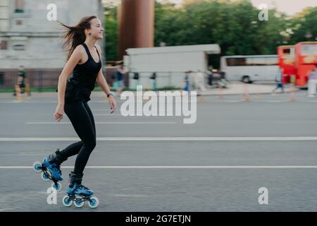 Energisch schlank junge lächelnde Frau Rollerblades auf der Straße der Stadt genießt die Freizeit aktiv bewegt sich schnell hat dunkle Haare auf Wind führt schweben Stockfoto