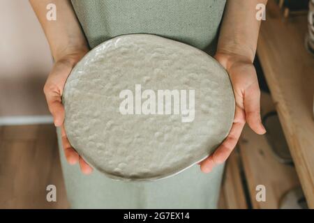 Eine Töpferin auf einer Schürze hält eine Keramikplatte in ihren Händen. Handgefertigt Stockfoto