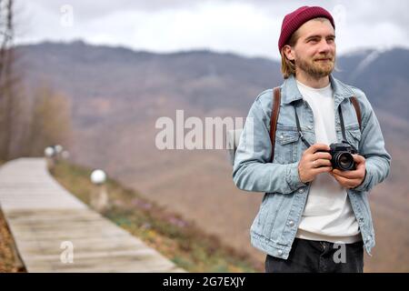 Fröhlicher bärtiger, männlicher Tourist mit Kamera, der unterwegs ist, Fotos macht, lächelt, zur Seite schaut, in lässigem Outfit. Speicherplatz kopieren. Tourismus, Fotografie, Reisen con Stockfoto