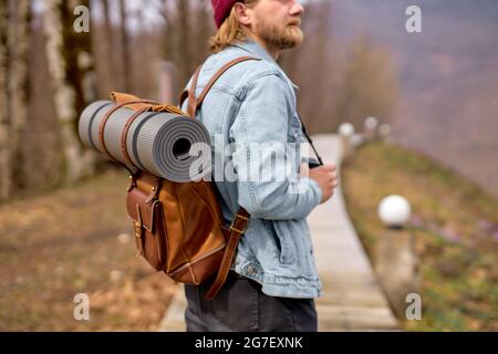 Hipster-Mann mit braunem Rucksack, der im Frühling die Natur erkundet und alleine im Freien unterwegs ist. Wanderer im lässigen Outfit, der in Wäldern unterwegs ist. Landschaft auf dem Land Stockfoto