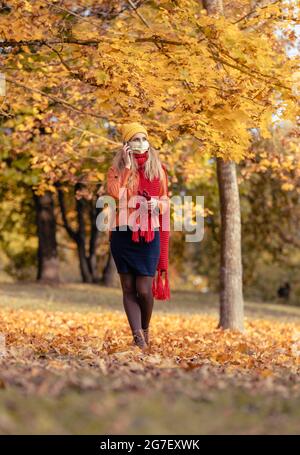 Frau mit ihrem Telefon zu Fuß in Herbst Park während Coronavirus-Krise inmitten buntem Laub Stockfoto