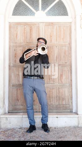 Jazzmusiker mit Brille und Bart, der auf der Straße posaunend steht. In englischer Mütze, Jacke und blauer Hose. Dahinter ist ein hölzernes d Stockfoto