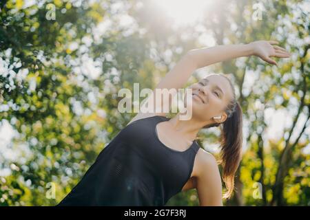 Sportliche Frau mit Pony Schwanz lehnt sich zur Seite Übungen im Freien gegen Bäume genießt sonnigen Tag hört Musik über kabellose Kopfhörer hat morgendliches Training Stockfoto