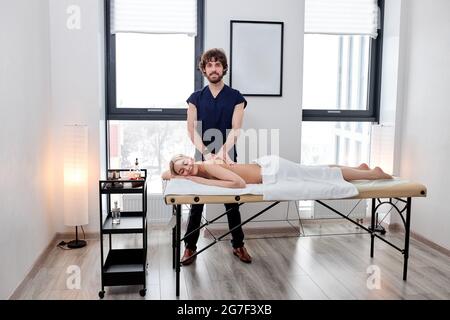 Portrait Of Bearded Nice Masseur In Uniform Sorgfältig Tun Massage Auf Dem Weiblichen Rücken, Blonde Kaukasische Frau Auf Bauch Liegend Entspannend, Genießen. Spa Kopieren Stockfoto