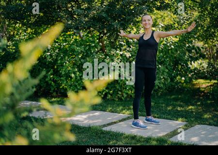 Outdoor-Schuss von athletischen fit junge Frau hält die Arme seitwärts in guter Laune Übungen im Freien trägt Sportlothes genießt frische Luft und schön Stockfoto