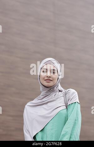 Porträt einer lächelnden, schönen jungen muslimischen Frau in Hijab und grünem Kleid, die gegen ein zeitgenössisches Gebäude steht Stockfoto