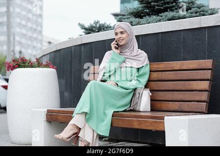 Lächelnd stilvolle junge muslimische Geschäftsfrau in langen grünen Kleid und Hijab sitzen auf der Bank mit Tasche und telefonieren Stockfoto