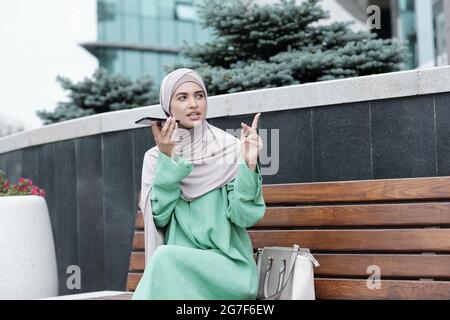 Junge muslimische Frau in religiösen Kleidern sitzt auf der Bank und zeigt mit dem Finger, während sie eine Audionachricht am Telefon aufzeichnet Stockfoto