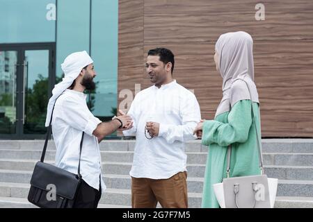 Zufriedene junge muslimische Geschäftspartner, die auf der Treppe gegen das Gebäude stehen und nach dem Treffen Handschlag geben Stockfoto