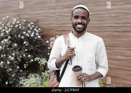 Porträt eines glücklichen jungen schwarzen Studenten Kerl in kufi Kappe hält Griff von Tasche und Kaffeetasse im Freien Stockfoto