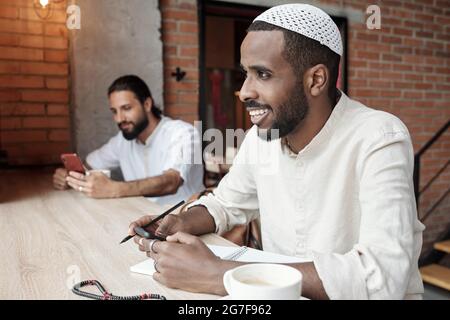 Lächelnder junger schwarzer Mann mit muslimischer Mütze sitzt am Tisch und schreibt Ideen auf Notizblock, während er eine Nachricht am Telefon schreibt Stockfoto