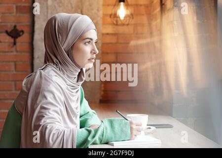 Ernsthafte nachdenkliche junge muslimische Frau im Hijab, die im Café am Tisch sitzt und Notizen in einem Skizzenblock macht Stockfoto