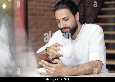 Lächelnder, hübscher junger bärtiger Mann in Ringen und Armbändern, der im Café am Tisch sitzt und Kaffee trinkt, während er sein Smartphone benutzt Stockfoto