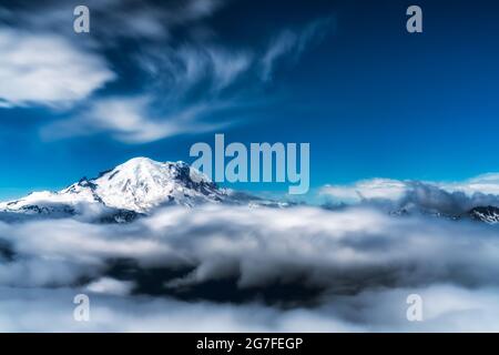 Mount Rainier in Washington hat seinen Kopf über den wispy Wolken unten. Dramatische Darstellung dieses Fotos mit tiefblauem Himmel und dunklem Peekaboo-Wald. Stockfoto