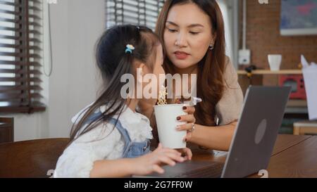 Asiatische Familie junge Mutter und ihre kleine Tochter lächeln mit Laptop-Computer zusammen für online lernen und Instant Nudeln zu Hause essen, Kind EA Stockfoto