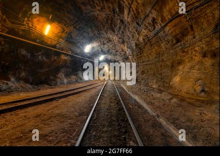 Tunnel des Bergbaus einer unterirdischen Mine. Viele Rohrleitungen an der Decke und Schienenbahn für Trolleys Stockfoto