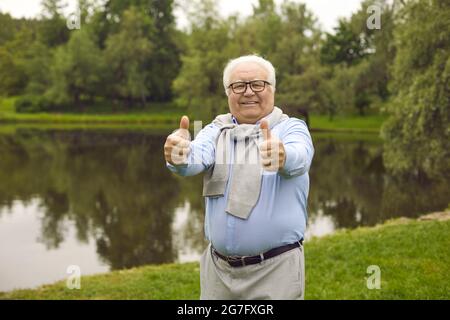 Porträt eines glücklichen Senioren, der in einem grünen Park steht und den Daumen nach oben gibt Stockfoto