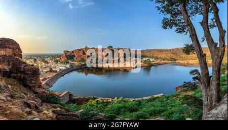 Agasthya Lake und die umliegenden roten Felshügel. Einige braune Steintempelgebäude verstreut herum, Badami, Karnataka, Indien. Stockfoto