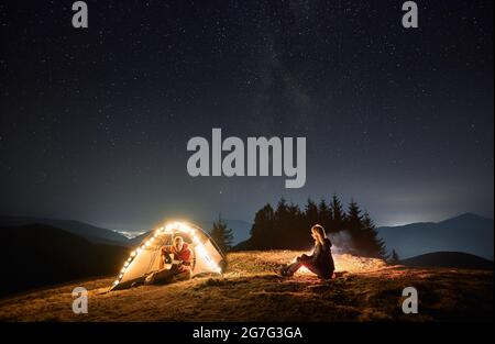 Junger Mann, der im beleuchteten Zeltlager sitzt und Gitarre spielt, während er Zeit mit seiner Freundin in den Bergen unter blauem Nachthimmel verbringt Stockfoto