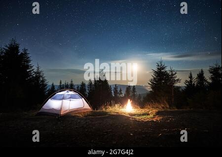 Weißes Touristenzelt auf einem Berghang in der Nähe eines brennenden Lagerfeuers am Wald. Mondschein im magischen Sternenhimmel Übernachtung in den Bergen. Co Stockfoto