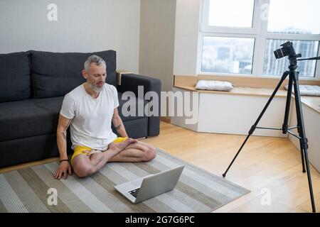 Ein reifer Mann in weißem T-Shirt sitzt auf dem Boden und zeichnet ein Online-Yoga-Tutorial auf Stockfoto