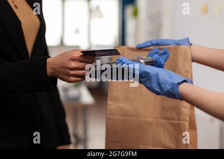 Nahaufnahme einer Unternehmerin, die eine Bestellung zum Essen zum Mitnehmen mit Kreditkarte über ein kontaktloses POS-Terminal im Startup-Büro bezahlt. Der Lieferer bringt die Lunchbox am Arbeitsplatz Stockfoto