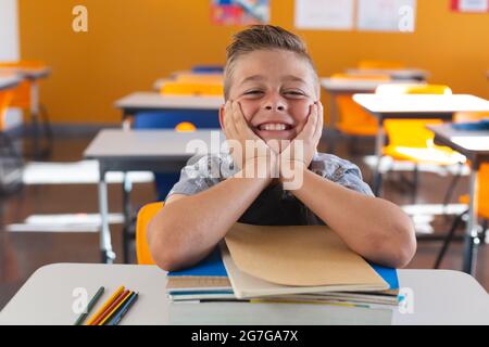 Fröhlicher kaukasischer Schuljunge, der im Klassenzimmer am Schreibtisch saß und sich an Büchern lehnte und lächelte Stockfoto