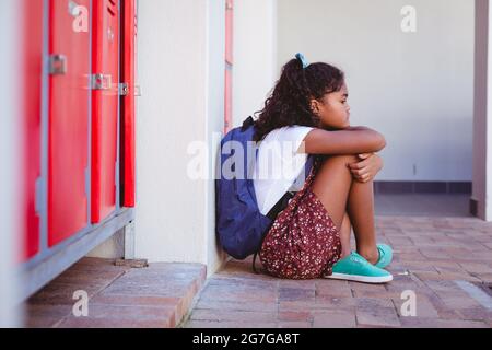 Unglückliches afroamerikanisches Schulmädchen, das an Schließfächern im Schulkorridor mit Schultasche sitzt Stockfoto