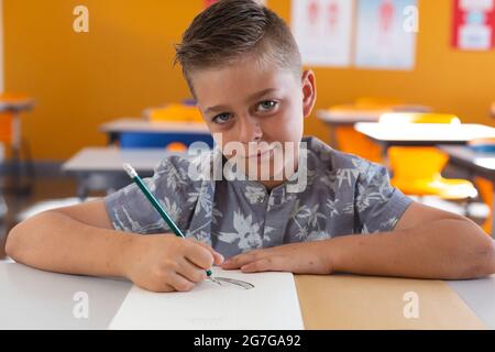 Porträt eines kaukasischen Schuljungen, der im Klassenzimmer am Schreibtisch sitzt, mit Bleistift im Buch und lächelnd gezeichnet Stockfoto