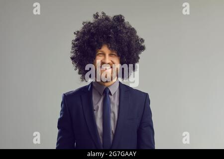 Studioportrait eines glücklich lächelnden jungen Geschäftsmannes in einem Anzug und einer lustigen lockigen Perücke Stockfoto