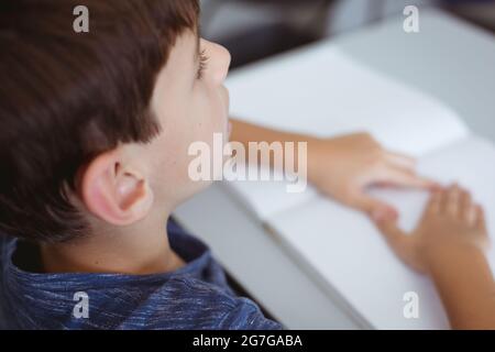 Blinder kaukasischer Schuljunge, der am Schreibtisch sitzt und mit den Fingern ein Braillebuch liest Stockfoto