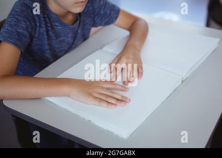 Blinder kaukasischer Schuljunge, der am Schreibtisch sitzt und mit den Fingern ein Braillebuch liest Stockfoto